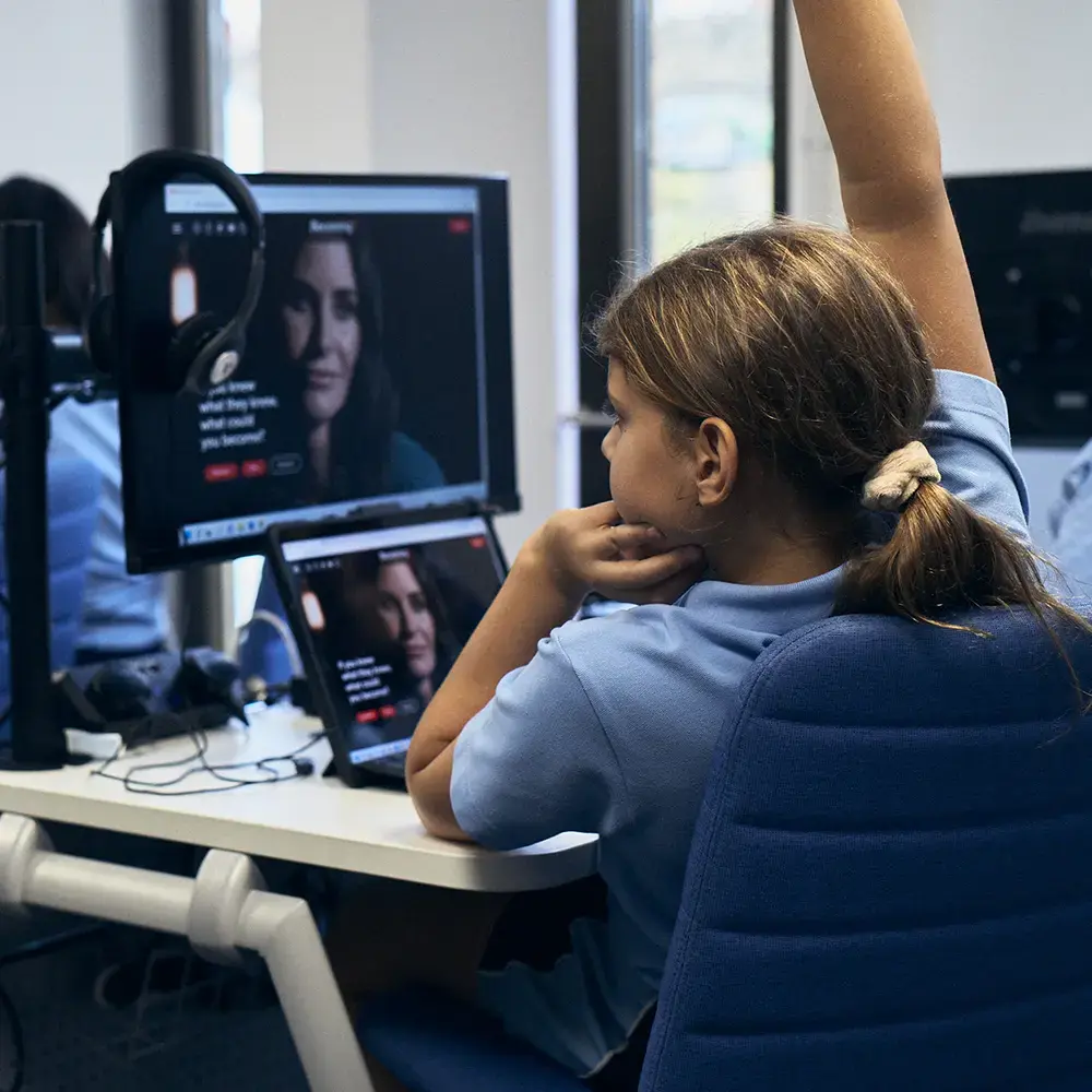 A student in a PSHE lesson with her hand up and the BecomingX Education platform on her computer screen.