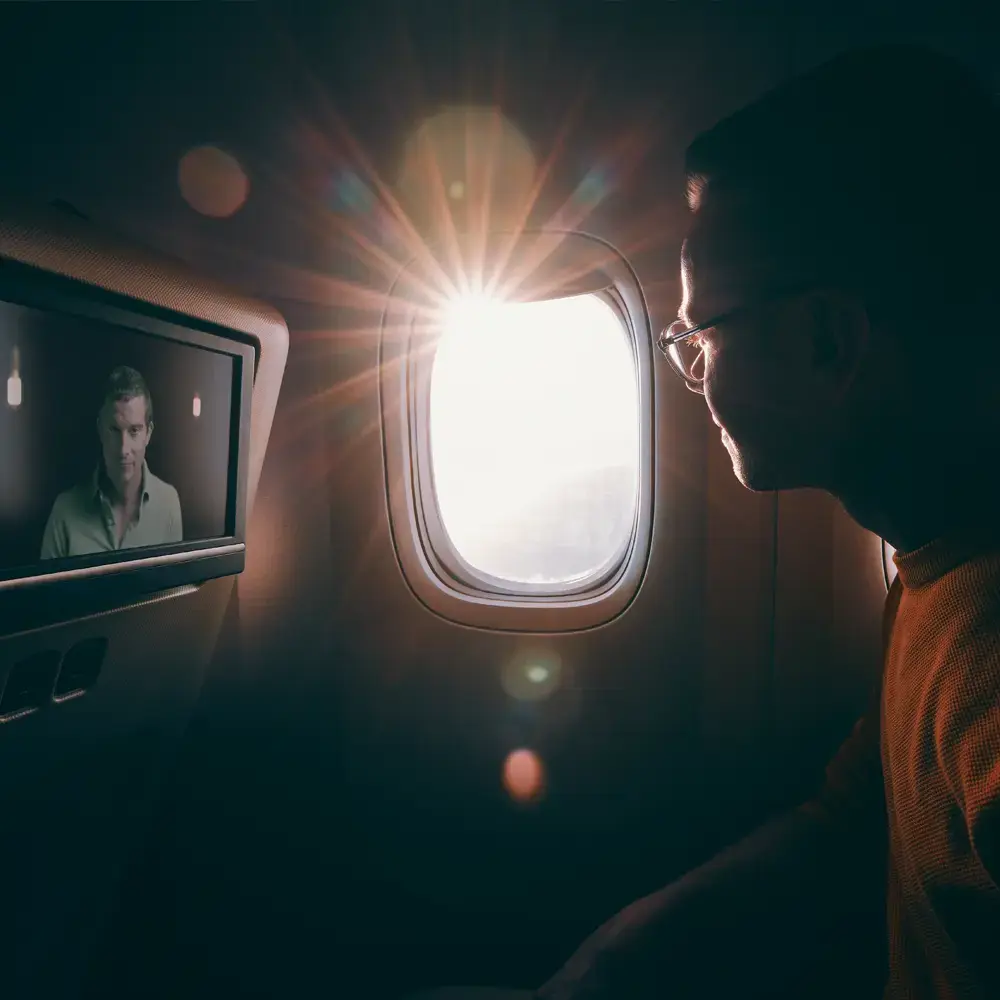 A man looking out an airplane window with a BecomingX film featuring Bear Grylls, playing on his screen.